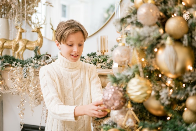 Chico feliz en suéter en el árbol de navidad decora y mira desde detrás tiene un tiempo feliz en c ...