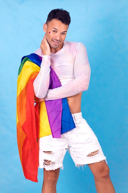 chico gay sonriendo con la bandera del orgullo sobre su hombro fondo azul