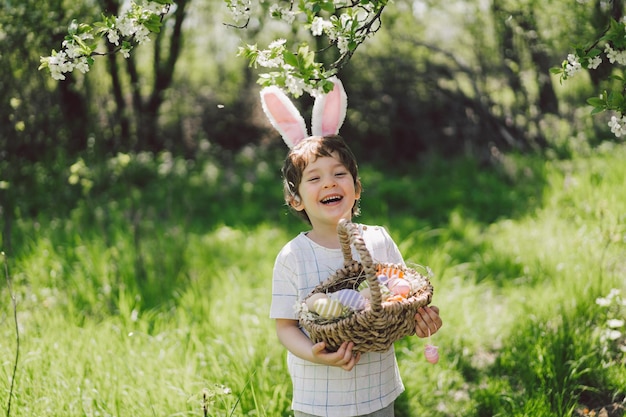 Chico gracioso con canasta de huevos y orejas de conejo en la caza de huevos de Pascua en el jardín soleado de primavera