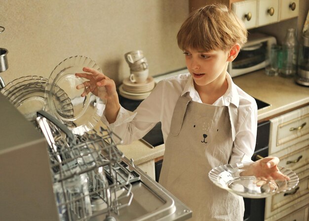 Un chico guapo de unos 10 años con un delantal descarga platos limpios del lavavajillas