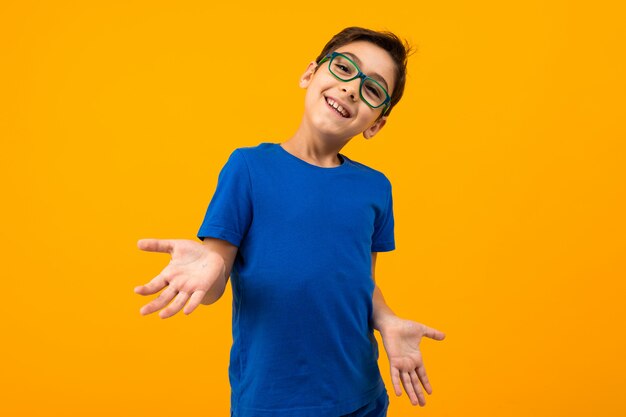 Chico guapo adolescente en una camiseta azul con gafas extiende sus manos sobre un fondo amarillo con espacio de copia