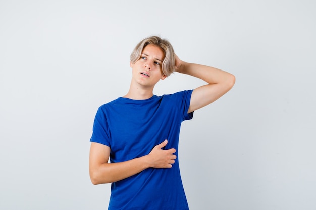 Chico guapo adolescente manteniendo la mano detrás de la cabeza, mirando a otro lado en camiseta azul y mirando pensativo, vista frontal.