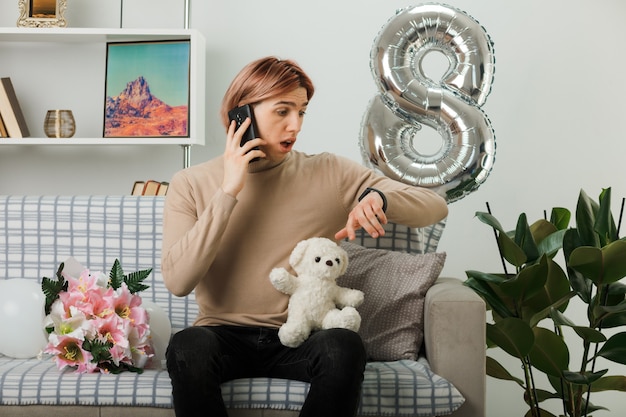 Foto chico guapo asustado en el día de la mujer feliz con oso de peluche habla por teléfono mirando el reloj de pulsera sentado en el sofá en la sala de estar