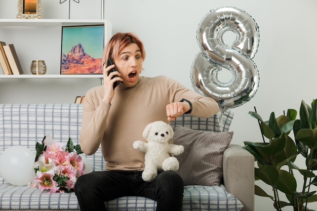 Chico guapo asustado en el día de la mujer feliz con oso de peluche habla por teléfono mirando el reloj de pulsera sentado en el sofá en la sala de estar