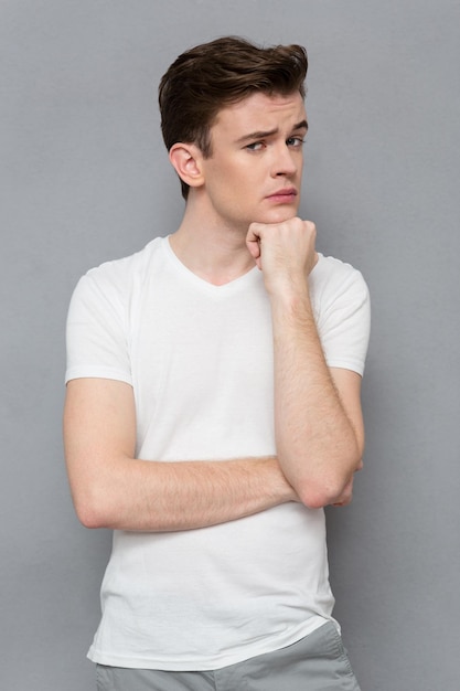 Chico guapo atractivo joven pensativo en camiseta blanca mirando, sosteniendo la mano en la barbilla y de pie contra la pared gris