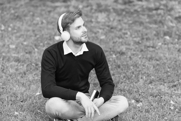 Un chico guapo con auriculares se relaja en el parque. El estudiante de tecnología de la vida moderna obtiene información del concepto de aprendizaje electrónico del libro. El hombre escucha música con auriculares y sostiene el libro.