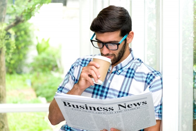 Chico guapo con barba y con gafas está tomando café y leyendo el periódico, de pie contra el pilar junto al balcón. Empresario con cara de serio.