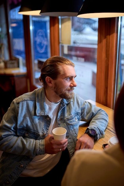 Chico guapo con barba tomando café en la cafetería, sonriendo, mirando al lado