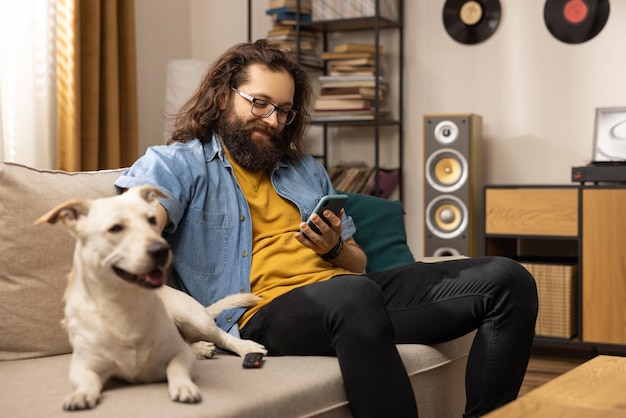 Un chico guapo barbudo está sentado en el sofá con su perro en la sala de estar, el hombre barbudo es br