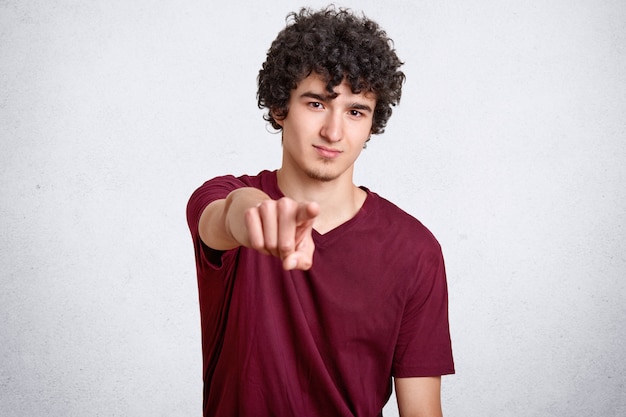 Chico guapo con cabello oscuro y rizado apunta directamente a la cámara con el dedo índice, expresa su elección, vestido con una camiseta informal