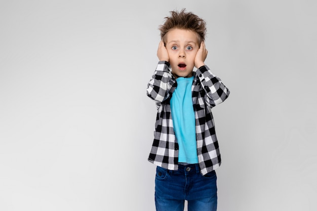 Foto un chico guapo con una camisa a cuadros, camisa azul y jeans se encuentra en un gris. el niño abrió la boca y se cubrió los oídos con las manos.