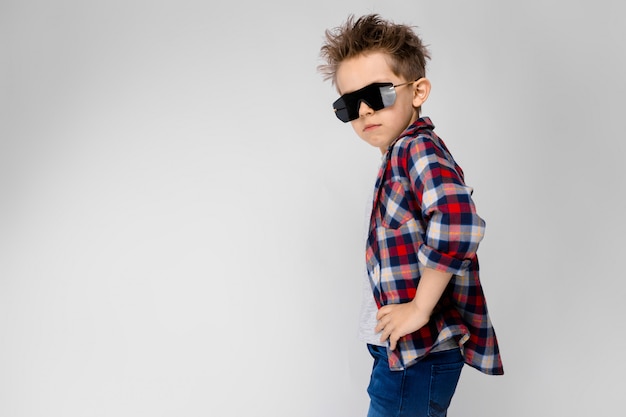 Un chico guapo con una camisa a cuadros, camisa gris y pantalones vaqueros se encuentra. El chico de las gafas de sol negras. El niño está medio despierto. El niño puso sus manos en sus caderas.