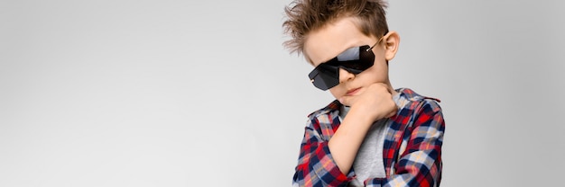 Un chico guapo con una camisa a cuadros, camisa gris y pantalones vaqueros se encuentra. El chico de las gafas de sol negras. El niño lleva una mano a la barbilla.