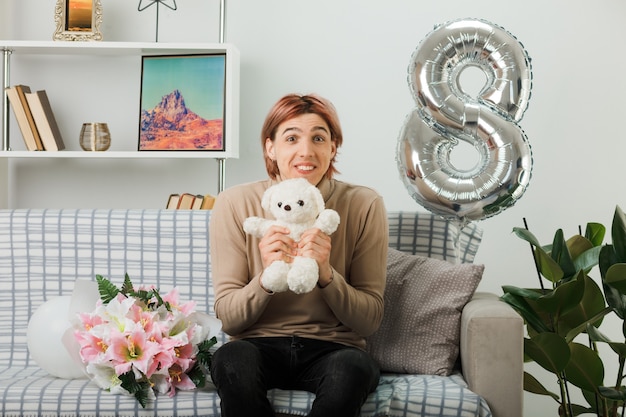 Chico guapo complacido en el día de la mujer feliz con oso de peluche sentado en el sofá en la sala de estar