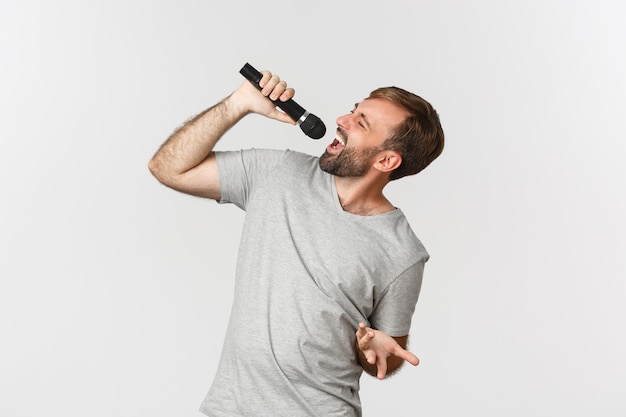 Foto chico guapo despreocupado cantando canciones en karaoke, sosteniendo el micrófono, de pie sobre fondo blanco.