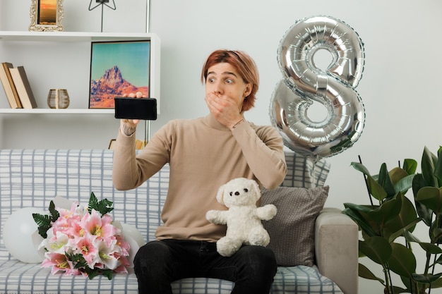 Foto chico guapo en el día de la mujer feliz con oso de peluche con teléfono sentado en el sofá en la sala de estar