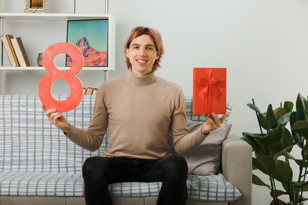 Chico guapo en el día de la mujer feliz sosteniendo el número ocho con presente sentado en el sofá en la sala de estar