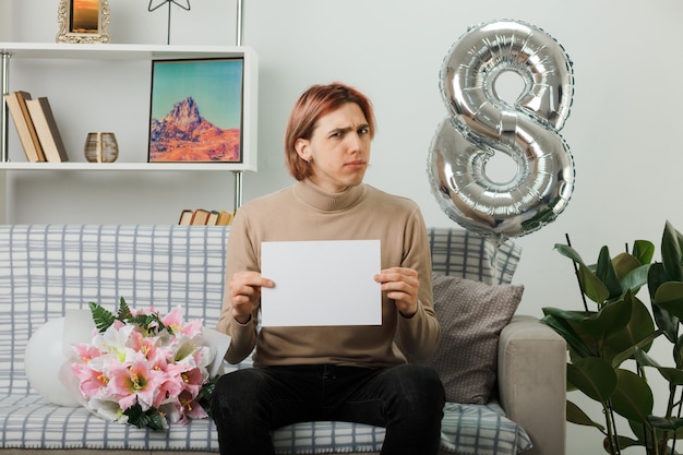 Chico guapo disgustado en el día de la mujer feliz celebración parpadeó sentado en el sofá en la sala de estar