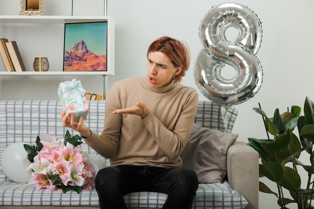 Chico guapo en feliz día de la mujer sosteniendo y puntos con mano presente sentado en el sofá en la sala de estar