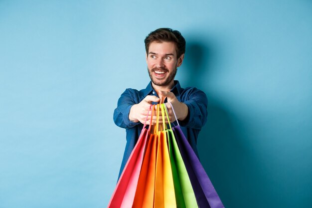 Chico guapo feliz sonriendo, estirar las manos con bolsas de la compra y mirando a un lado el espacio vacío, de pie sobre fondo azul.