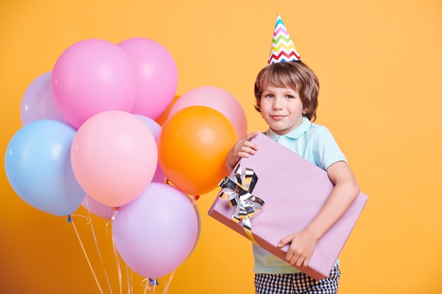 Chico guapo con gorro de cumpleaños posando con gran caja azul y globos de colores sobre pared amarilla