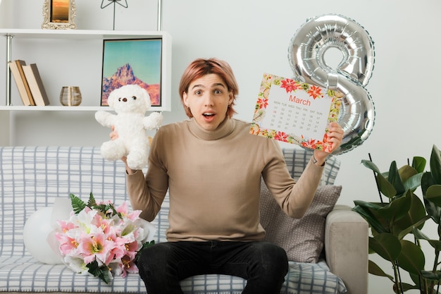 Chico guapo impresionado en el día de la mujer feliz con oso de peluche con calendario sentado en el sofá en la sala de estar