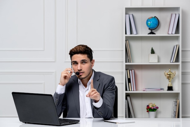 Chico guapo inteligente de servicio al cliente con una computadora portátil en un blazer de oficina gris que muestra un gesto de espera