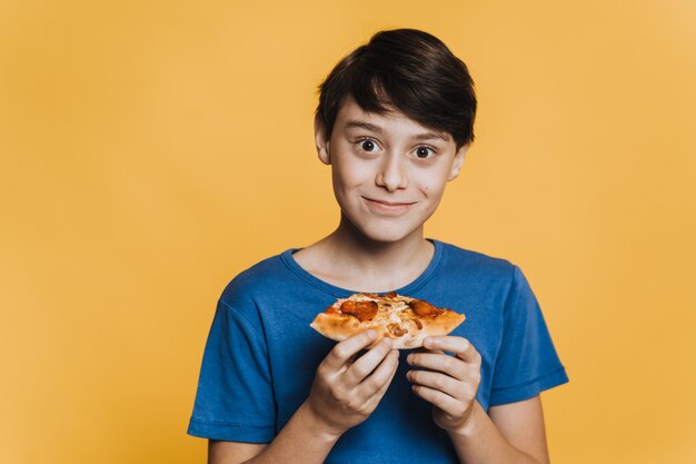 Chico guapo joven en camiseta azul con los ojos bien abiertos, muy feliz porque consiguió su pizza en casa, de pie sobre fondo amarillo. Quedarse en casa