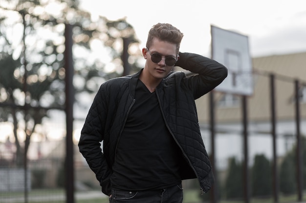 Chico guapo joven con estilo hipster con gafas de sol de moda en ropa negra de moda en la calle