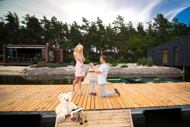 Chico guapo joven hace una propuesta de matrimonio a su amada niña, de pie sobre sus rodillas en un muelle de madera. Romance y amor en un muelle de madera