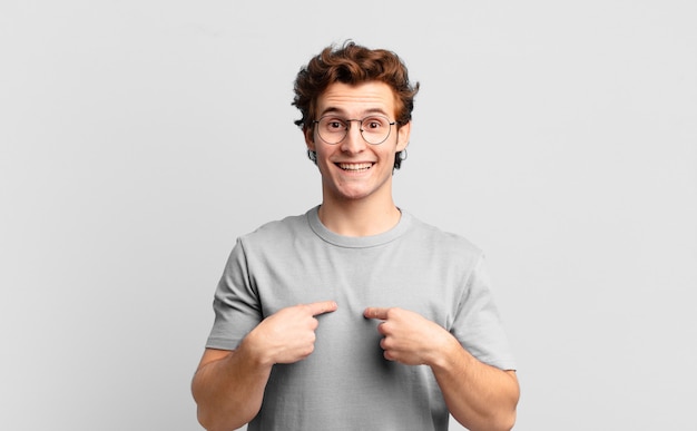 Chico guapo joven que se siente feliz, sorprendido y orgulloso, señalando a sí mismo con una mirada emocionada y asombrada