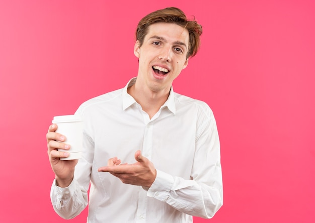 Chico guapo joven sonriente con camisa blanca sosteniendo y puntos con la mano en la taza de café aislado en la pared rosa