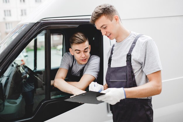 Un chico guapo joven vestido con uniforme está apuntando al portapapeles mientras está sentado en el coche. Otro trabajador con uniforme está mirando el portapapeles. Mudanza de casa, servicio de mudanza.