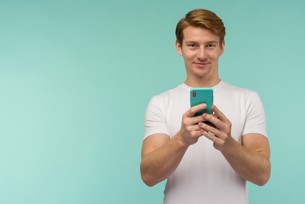 Chico guapo pelirrojo deportivo con camiseta blanca toma un selfie o transmite en línea en un teléfono inteligente sobre un fondo azul. - imagen