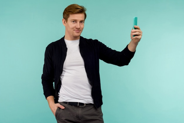 Chico guapo pelirrojo deportivo toma una selfie o transmite en línea en un teléfono inteligente en un espacio azul.