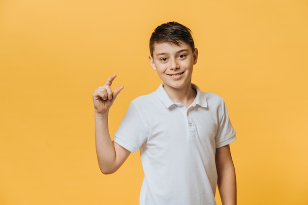 Chico guapo positivo con elegante peinado, vestido con una camiseta blanca, muestra algo muy pequeño o pequeño, de buen humor, posa contra la pared amarilla.