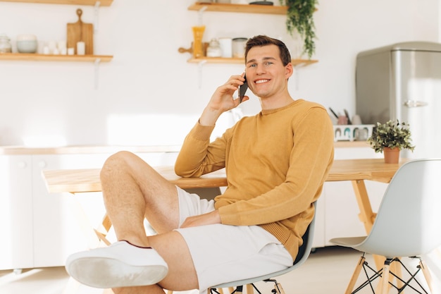 Chico guapo con ropa informal amarilla sentado en casa en la cocina tomando café y trabajando en tableta y teléfono