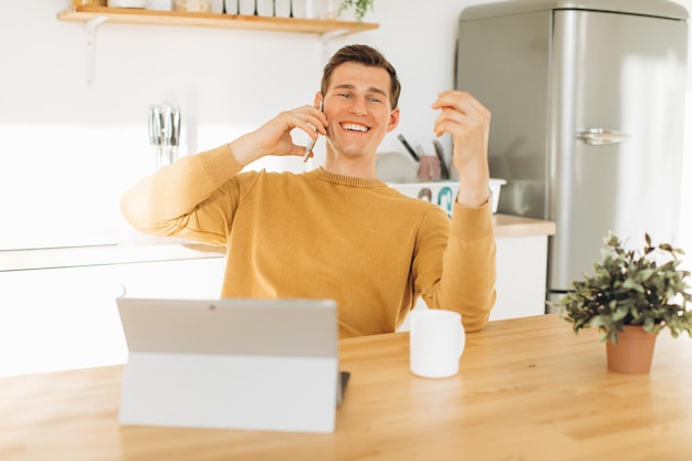 Chico guapo con ropa informal amarilla sentado en casa en la cocina tomando café y trabajando en tableta y teléfono