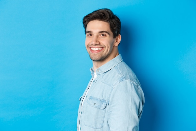 Chico guapo y seguro gira la cabeza al frente, sonriendo feliz, de pie sobre la pared azul