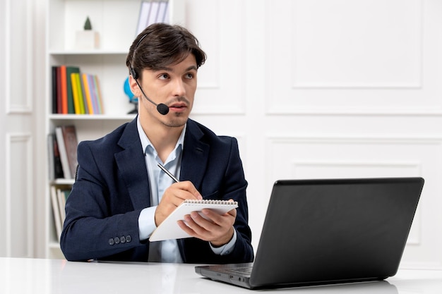 Chico guapo de servicio al cliente con auriculares y computadora portátil en traje tomando notas y mirando hacia arriba