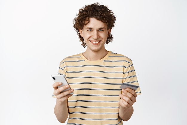 Chico guapo sonriente sosteniendo el teléfono móvil y el concepto de tarjeta de crédito de orden de compra en línea en el fondo blanco de la aplicación