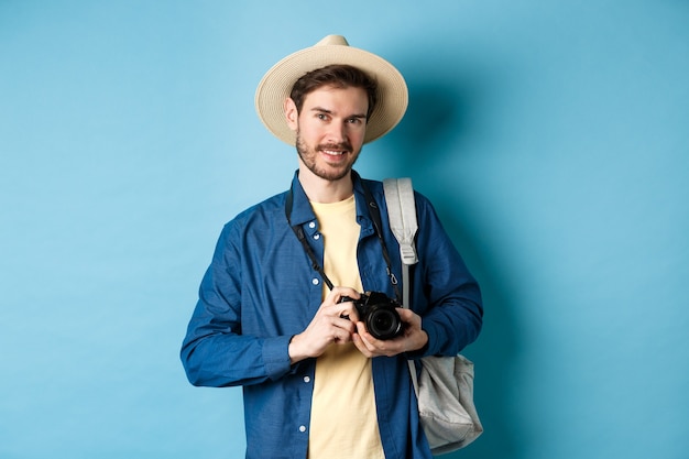 Chico guapo va de vacaciones de verano, mochilero de vacaciones. Turista con sombrero de paja y cámara sonriendo feliz, de pie sobre fondo azul.