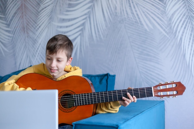 Chico guapo vestido con jeans casuales y sudadera amarilla ve una lección de video sobre tocar la guitarra en el acogedor sofá de casa