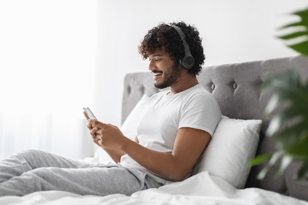 Chico hindú alegre usando teléfono celular y auriculares en la cama