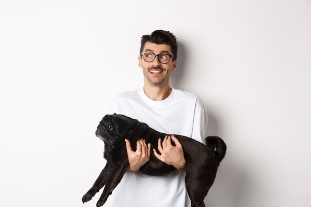 Chico hipster emocionado con su perro mirando a la izquierda en el logo. El dueño de una mascota guapo lleva pug negro y mira fijamente la pancarta promocional, de pie sobre el fondo blanco.