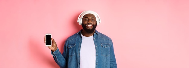 Chico hipster feliz escuchando música en los auriculares y mostrando la pantalla del móvil sonriendo satisfecho de pie