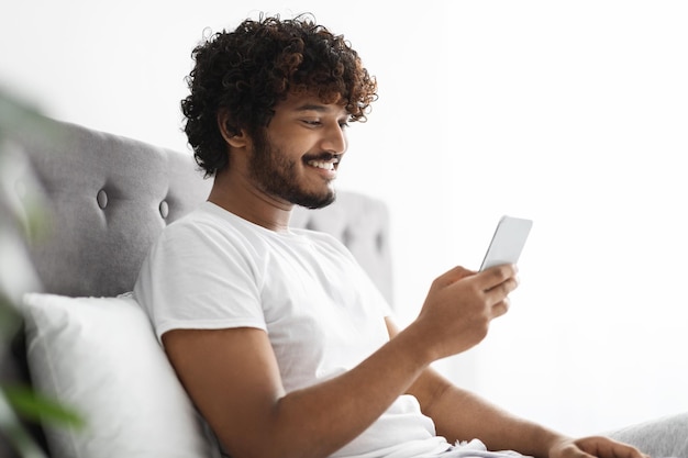 Chico indio positivo desplazándose en la cama usando un teléfono inteligente