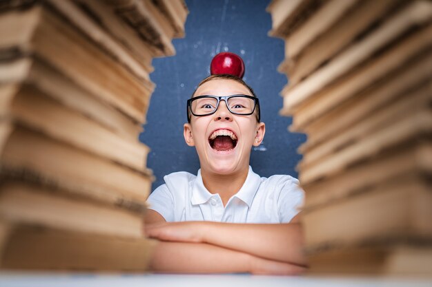 Foto chico inteligente feliz en vasos sentado entre dos pilas de libros con manzana roja en la cabeza y mira a la cámara sonriendo con la boca abierta.