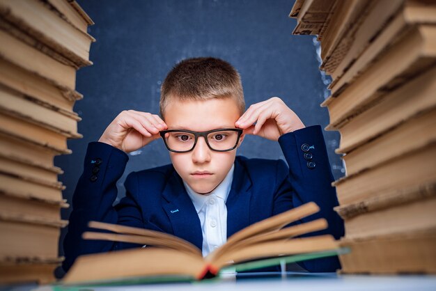 Chico inteligente con gafas sentado entre dos pilas de libros y leer el libro pensativamente.