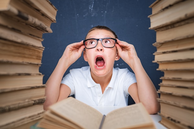 Chico inteligente con gafas volviéndose loco por leer un libro mientras está sentado entre dos pilas de libros